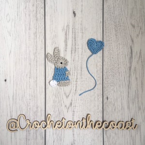 May include: A crocheted bunny wearing a blue dress and a white tail, next to a blue crocheted heart balloon on a white wood background. The text "@CrochetonTheCoast" is written in wood letters below the bunny and balloon.