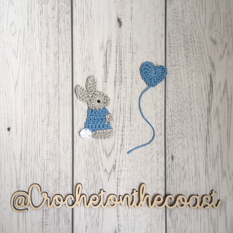 May include: A crocheted bunny wearing a blue dress and a white tail, next to a blue crocheted heart balloon on a white wood background. The text "@CrochetonTheCoast" is written in wood letters below the bunny and balloon.