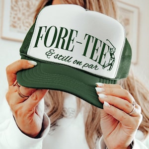 May include: A green and white trucker hat with the text "FORE-TEE" and "& still on par" in green. The hat features a golf-themed graphic of a golfer swinging a club. The hat has a green brim and mesh back.