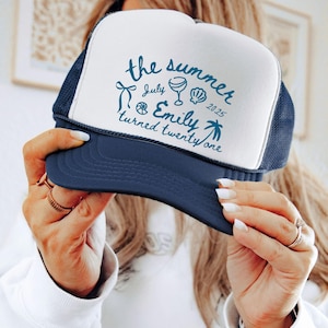 May include: A navy blue and white trucker hat with the text "the summer July Emily turned twenty one 2025" in teal. The hat has a mesh back and a curved peak. The text includes illustrations of a bow, a wine glass, a seashell, and a palm tree.