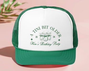 May include: A white and green trucker hat with the text "A Tini Bit Older" and "Kim's Birthday Party" in green. The hat has a mesh back and a green brim.
