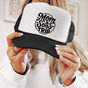 May include: A black and white trucker hat with the text "Mom & Sons TRIP 2026 CABO" in a circular design. The hat has a black brim and mesh back, and a white front panel.