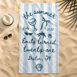 May include: A rectangular beach towel with light blue and white vertical stripes, featuring dark blue text that reads the summer July 2025 Emily turned twenty-one Destin, Fl, alongside illustrations of a cocktail glass, bow, sunglasses, palm tree, and shells.