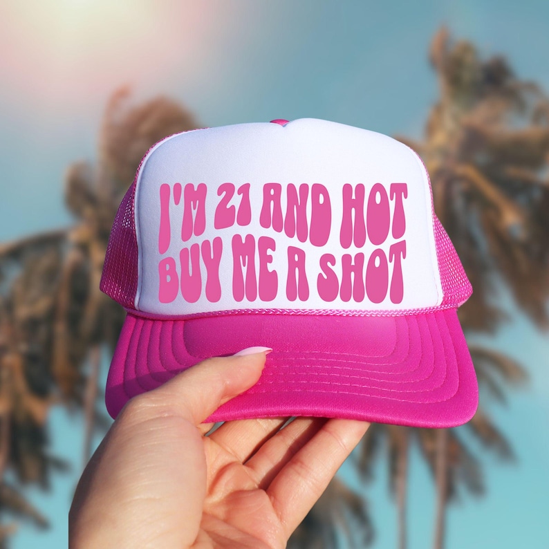 May include: A pink and white trucker hat with the text "I'M 21 AND HOT BUY ME A SHOT" in pink. The hat has a pink brim and mesh back, and is held up against a blurred tropical background.
