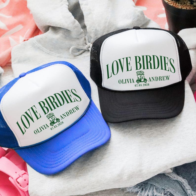 Puede incluir: Dos gorras de camionero con el texto "LOVE BIRDIES" en verde. Una gorra es azul con la parte delantera blanca, y la otra es negra con la parte delantera blanca. Ambas gorras tienen los nombres "OLIVIA ANDREW" y la fecha "07.03.2026".