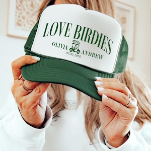 Puede incluir: Gorra de camionero verde y blanca con el texto "LOVE BIRDIES" en verde. La gorra tambi&eacute;n incluye los nombres "OLIVIA ANDREW" y la fecha "07.03.2026" en verde. La gorra tiene una visera verde y una parte trasera de malla.
