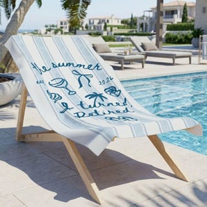 Pu&ograve; includere: Un telo mare a righe azzurre e bianche drappeggiato su una sedia a sdraio in legno. Il telo presenta una scritta blu scuro che recita the summer est 2026 I turned Retired, accompagnata da illustrazioni di un calice, palma e infradito.