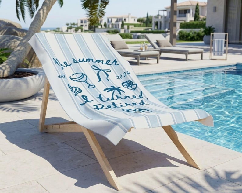 Pu&ograve; includere: Un telo mare a righe azzurre e bianche drappeggiato su una sedia a sdraio in legno. Il telo presenta una scritta blu scuro che recita the summer est 2026 I turned Retired, accompagnata da illustrazioni di un calice, palma e infradito.