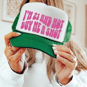 May include: Green and white trucker hat with the text "I'M 21 AND HOT BUY ME A SHOT" in pink. The hat has a green brim and mesh back, and a white front panel.