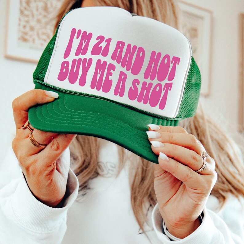 May include: Green and white trucker hat with the text "I'M 21 AND HOT BUY ME A SHOT" in pink. The hat has a green brim and mesh back, and a white front panel.