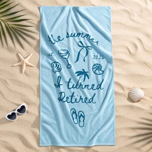 Pu&ograve; includere: Un telo mare rettangolare azzurro con testo blu scuro che recita the summer est. 2026 I turned Retired, accompagnato da illustrazioni grafiche di un calice da vino, un fiocco, una palma, conchiglie e un paio di infradito.