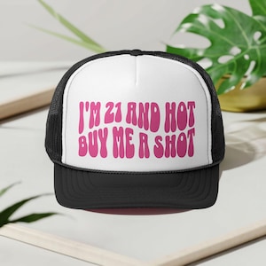May include: Black and white trucker hat with a white front panel. The front of the hat features the text "I'M 21 AND HOT BUY ME A SHOT" in pink, retro-style lettering. The hat has a curved brim and mesh back.