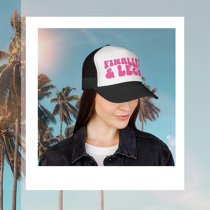 May include: Black and white trucker hat with the text "FINALLY & LEGALLY" in pink. The hat has a mesh back and a curved brim. The hat is worn by a person in front of a tropical background.
