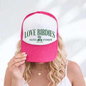 Puede incluir: Gorra de camionero rosa y blanca con el texto "LOVE BIRDIES" en verde. La gorra presenta los nombres "Olivia" y "Andrew" con un gr&aacute;fico de carrito de golf y la fecha "07.03.2026".