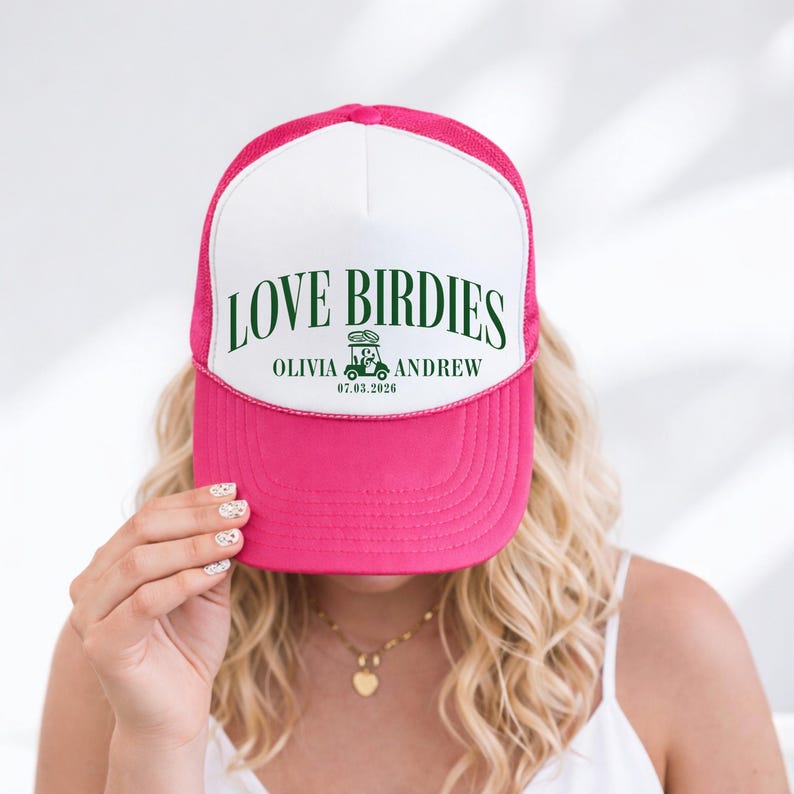 Puede incluir: Gorra de camionero rosa y blanca con el texto "LOVE BIRDIES" en verde. La gorra presenta los nombres "Olivia" y "Andrew" con un gr&aacute;fico de carrito de golf y la fecha "07.03.2026".