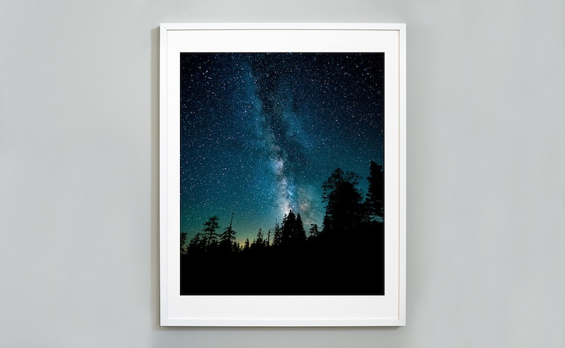 May include: A framed photograph of a dark blue night sky with a bright white Milky Way galaxy visible. The silhouette of a forest of trees is in the foreground.