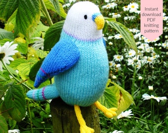 May include: A knitted blue and white parakeet with a yellow beak and feet sits on a wooden post. The bird has blue wings and a striped tail. The background includes green leaves and white flowers. Text on the right says "Instant download PDF knitting pattern."