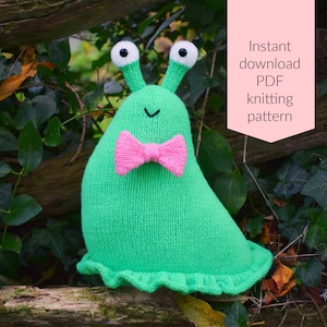 May include: A green knitted slug with a pink bow tie and white eyes. The slug is sitting on a wooden branch with green leaves.