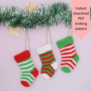May include: Three miniature knitted Christmas stockings in red, green, and white stripes hang from a green garland with gold star decorations. The stockings have red, white, and green accents. Text on a pink banner reads "Instant download PDF knitting pattern."