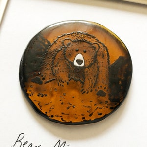 Puede incluir: Un adorno redondo de vidrio color ámbar con una ilustración en blanco y negro de un oso. El texto "Bear Minimum" está escrito debajo del adorno.