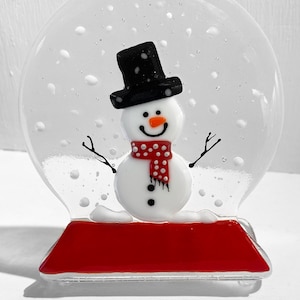 May include: A glass snow globe with a white snowman wearing a black top hat and a red and white polka dot scarf. The snowman is standing on a red base.