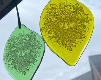 May include: Two glass ornaments shaped like leaves, each with a green man design. The left ornament is green and the right ornament is yellow.