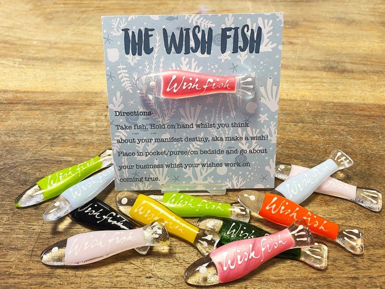 May include: A collection of colorful glass wish fish, each with the words "Wish Fish" written on them. The fish are arranged on a wooden surface with a card that reads "The Wish Fish" and instructions on how to use them.