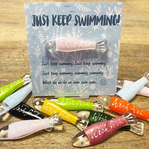 May include: A set of colorful glass fish ornaments with the words "Wish Fish" written on them. The ornaments are arranged on a wooden surface with a card that reads "Just Keep Swimming!"
