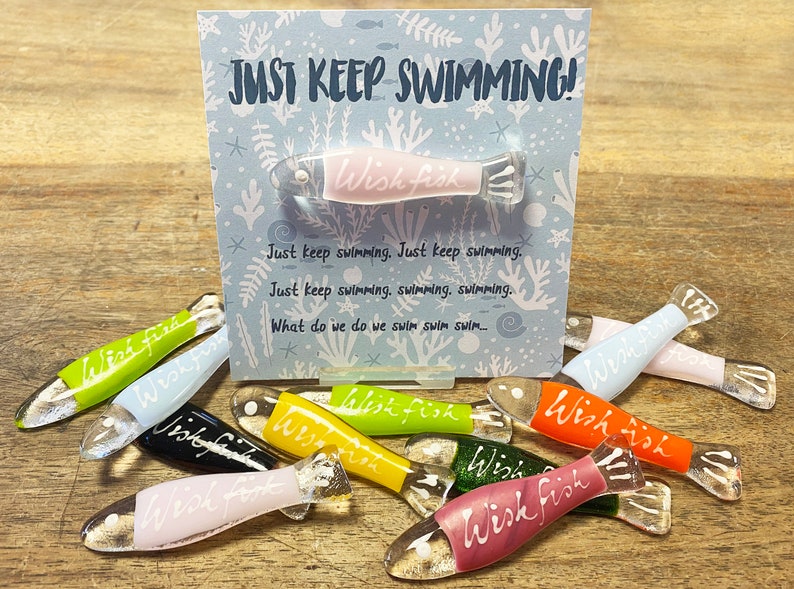 May include: A set of colorful glass fish ornaments with the words "Wish Fish" written on them. The ornaments are arranged on a wooden surface with a card that reads "Just Keep Swimming!"
