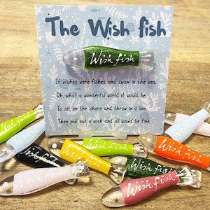 May include: A set of colorful glass wish fish ornaments with the text "Wish Fish" written on each one. The ornaments are displayed on a wooden surface with a card that reads "The Wish Fish" and a poem about wishing.
