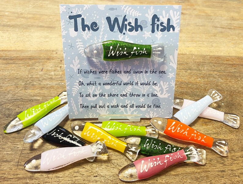 May include: A set of colorful glass wish fish ornaments with the text "Wish Fish" written on each one. The ornaments are displayed on a wooden surface with a card that reads "The Wish Fish" and a poem about wishing.