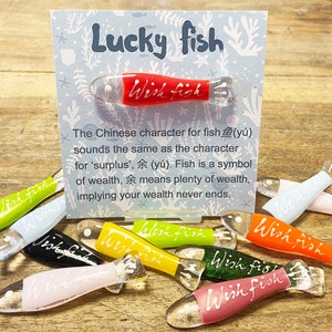 May include: A collection of colorful glass fish ornaments with the words "Wish Fish" written on them. The ornaments are arranged on a wooden surface with a card that reads "Lucky Fish" and explains the symbolism of the fish in Chinese culture.