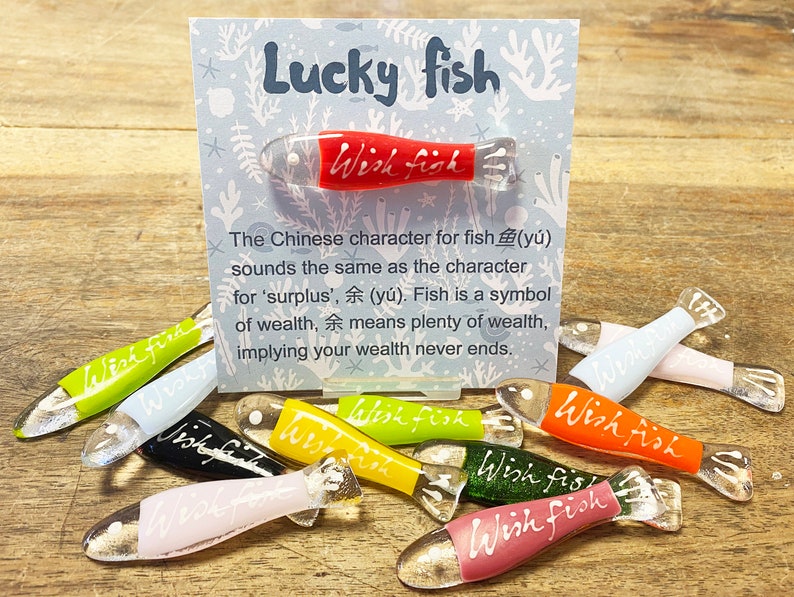 May include: A collection of colorful glass fish ornaments with the words "Wish Fish" written on them. The ornaments are arranged on a wooden surface with a card that reads "Lucky Fish" and explains the symbolism of the fish in Chinese culture.