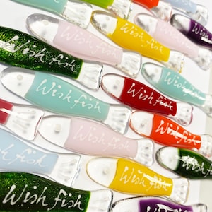May include: A collection of colorful glass fish ornaments with the words "Wish Fish" written on them. The fish are in various colors, including green, blue, pink, yellow, red, and purple.