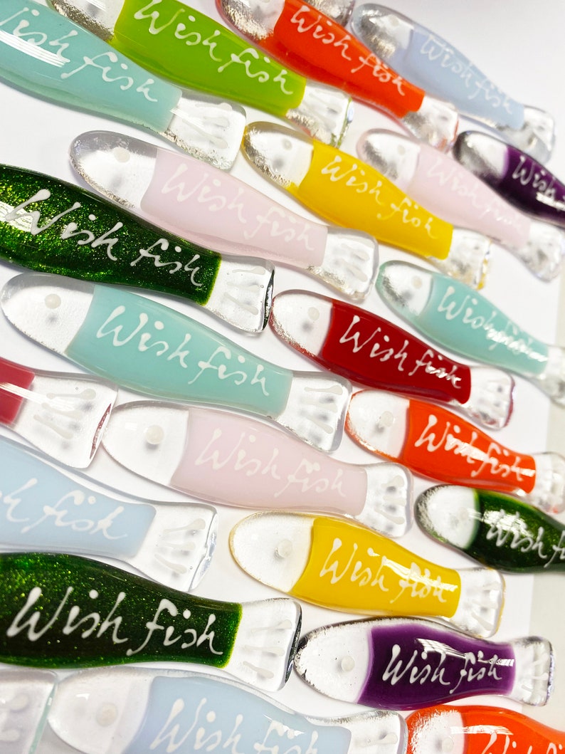 May include: A collection of colorful glass fish ornaments with the words "Wish Fish" written on them. The fish are in various colors, including green, blue, pink, yellow, red, and purple.