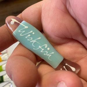 May include: A light blue glass fish with the word "wish fish" written in white on the side. The fish has a white dot on the top and two white dots on the tail.