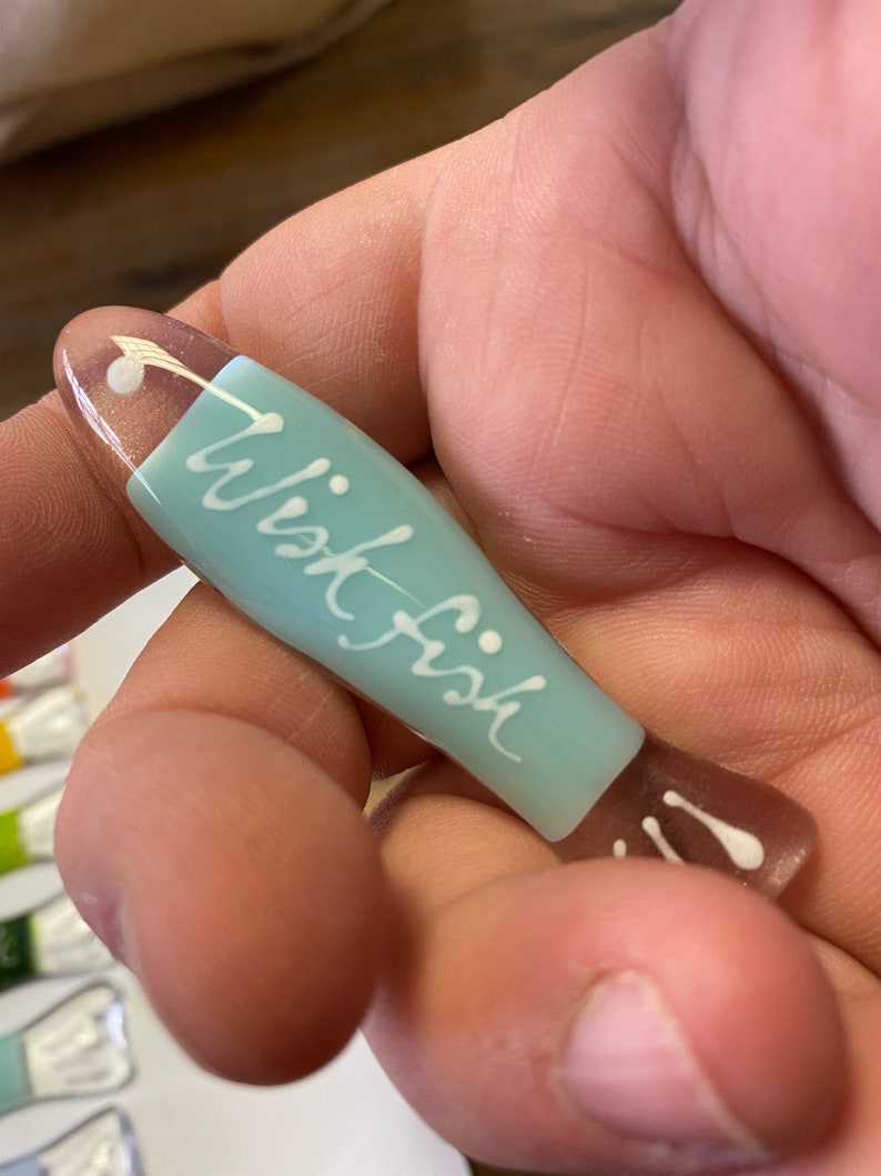 May include: A light blue glass fish with the word "wish fish" written in white on the side. The fish has a white dot on the top and two white dots on the tail.