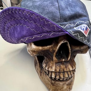 May include: A denim-style cap with a purple brim and white stitching, resting on a skull-shaped decorative object. The cap has a "MADE IN USA" patch. The skull is a light brown colour.