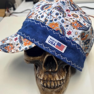 May include: A fabric cap featuring a white base with a colorful orange, blue, and red paisley pattern. The cap has a solid dark blue band across the front with white zigzag stitching and a rectangular white label that reads MADE IN USA with a flag.