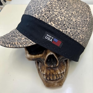 May include: A beige and black patterned cap with a black brim and band. The cap is labeled "MADE IN USA" with a small American flag. The cap is resting on a skull.