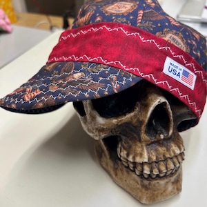 Może przedstawiać: Granatowa czapka z powtarzającym się wzorem bielików amerykańskich, gwiazd i napisem America's 250th Birthday. Czapka posiada szeroki, czerwony pas materiału z przodu z białymi szwami zygzakowatymi oraz białą metkę z napisem Made in USA.