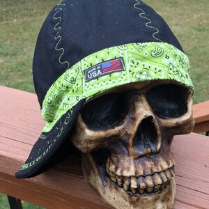 Puede incluir: Casquete de soldadura negro con ribete de bandana verde y una etiqueta "Made in USA". La gorra se lleva en una figura de calavera.