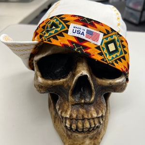 May include: White bandana with a colorful geometric pattern and the text "Made in USA" on a yellow label. The bandana is on a skull.