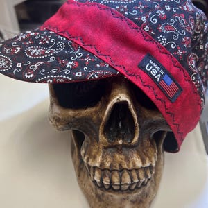 May include: A black and red paisley print cap with a red brim, resting on a skull. The cap has a small tag that reads "MADE IN USA" with an American flag.