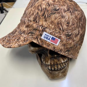 Może przedstawiać: Brązowo-czarna czapka z krótkim daszkiem, z białym szwem. Widoczna jest mała metka z napisem "MADE IN USA" i amerykańską flagą. Czapka spoczywa na czaszce.