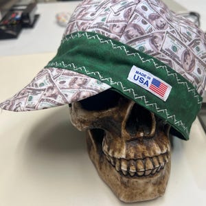 Pode incluir: Um boné de tecido com um padrão repetitivo de notas de cem dólares. O boné tem uma faixa larga verde-escura na frente com costuras em ziguezague brancas. Uma pequena etiqueta retangular branca traz a inscrição MADE IN USA com uma bandeira.
