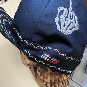 Puede incluir: Gorra de béisbol negra con una mano de esqueleto bordada en blanco haciendo el dedo medio. La gorra tiene una puntada en zigzag blanca alrededor del borde y una etiqueta "Made in USA" con una pequeña bandera estadounidense.