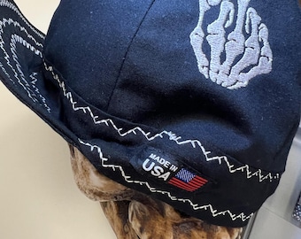 May include: Black baseball cap with a white embroidered skeleton hand giving the middle finger. The cap has a white zig-zag stitch around the brim and a "Made in USA" label with a small American flag.