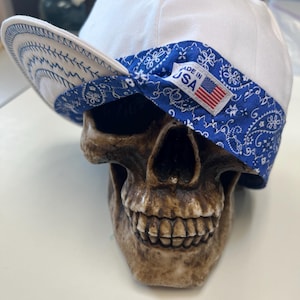 May include: A white welder's cap with a blue paisley patterned band and brim, resting on a skull. The cap has a small tag that reads "MADE IN USA" with an American flag. The brim is decorated with white stitching.