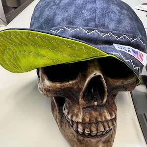 Puede incluir: Una gorra de soldadura azul oscuro y verde con un borde verde contrastante y costuras blancas. La gorra tiene una etiqueta "Made in USA" en el lateral.